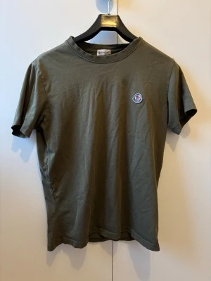 Olivgrön Moncler t-shirt i bomull - Snygg olivgrön t-shirt från Moncler med klassisk passform och rund hals. Tillverkad i mjuk bomull och har Monclers ikoniska logga broderad på bröstet. Perfekt basplagg för en clean och stilren look. Passar även storlek S! 