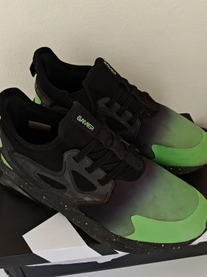 Svart-gröna sneakers från Gamer 37,5 - Snygga sneakers från Gamer i svart och grönt med cool gradient-effekt på ovandelen. Skorna har svarta snören, svart sula med vita prickar och detaljer i plast längs sidorna. Ovandelen är i textil och syntet, perfekt för en streetwear-look. Storlek 37,5 / 38 . Använda måttligt men fortfarande i ny skick. Ny pris : 40 till 60 EURO .