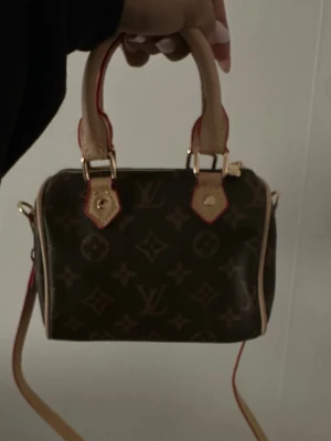 monogram handväska - Säljer en klassisk brun handväska  med ikoniskt monogrammönster. Väskan har beige handtag, guldfärgade detaljer och en avtagbar axelrem. Tillverkad i canvas med läderdetaljer och röda kanter för en lyxig touch.