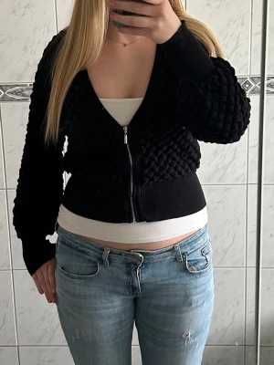  zip-kofta  - Säljer en svart kofta med dragkedja med coolt strukturmönster.  Koftan har ballongärmar och breda muddar vid ärmslut och midja. Storlek M men passar xs-m. 