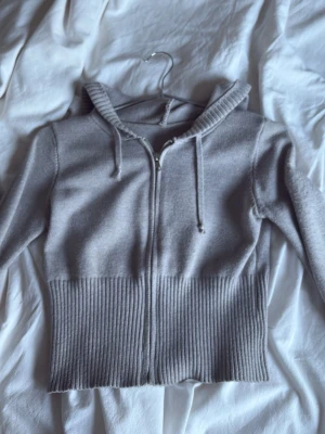 Grå zip hoodie med ribbade detaljer - Säljer en stilren grå hoodie med dragkedja och huva. Hoodien har ribbade muddar och bred ribbad nederkant som ger en snygg passform. Tillverkad i mjukt stickat material och har dragsko i huvan. Perfekt för en avslappnad look.