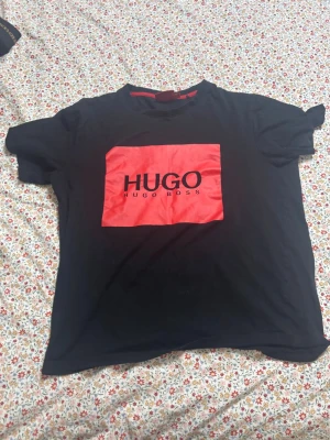 Svart HUGO BOSS t-shirt med röd logga - Snygg svart t-shirt från HUGO BOSS med stor röd ruta och svart logotyp på bröstet. Klassisk passform och rund halsringning. Perfekt för dig som gillar stilrena plagg med tydlig branding.
