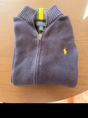 Mörkgrå stickad tröja Polo Ralph Lauren - Snygg mörkgrå stickad tröja från Polo Ralph Lauren med dragkedja och gul broderad logga på bröstet. Tröjan har hög krage och ribbstickade detaljer. Perfekt för dig som gillar klassisk och stilren design. Orginal boxen ingår 