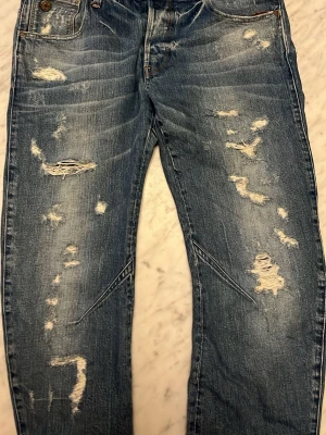 Blå slitna jeans med hål - Säljer ett par blå jeans med distressed detaljer och flera hål framtill för en cool, avslappnad look. Jeansen har klassisk femficksdesign och raka ben. Materialet är denim i bomull. Perfekta för dig som gillar streetwear och en trendig stil.
