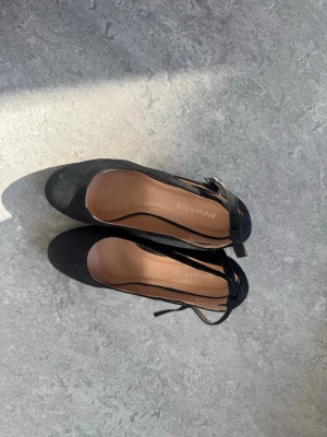 Svarta pumps med ankelrem från Anna Field - Snygga svarta pumps från Anna Field med rund tå och ankelrem med spänne. Skorna har en klassisk klack och är tillverkade i mockaimitation med beige innersula. Perfekta för dig som vill ha en stilren och tidlös look.