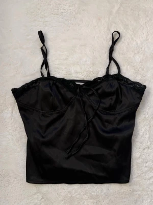 Svart satinlinnetopp från Venderby's - Snygg svart linnetopp från Venderby's i glansigt satinmaterial. Toppen har tunna axelband, spetsdetaljer längs urringningen och en liten knytning framtill. Perfekt för att lyfta vilken outfit som helst.
