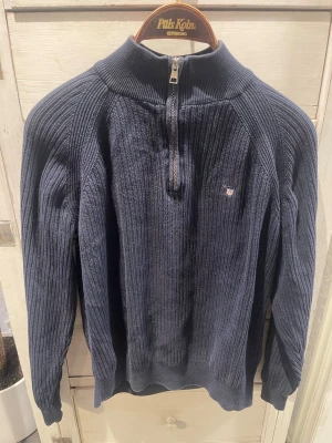 Mörkblå stickad halvzip från Gant - Mörkblå stickad tröja från Gant med halvzip och hög krage. Tröjan har ribbstickad struktur, lång ärm och en liten broderad Gant-logga på bröstet. Perfekt för lager på lager och en klassisk stil.