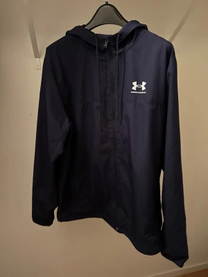 Marinblå vindjacka från Under Armour - Snygg marinblå vindjacka från Under Armour med huva och dragkedja framtill. Jackan har meshfoder på insidan och en vit logga på bröstet. Perfekt för blåsiga dagar och sportiga outfits.