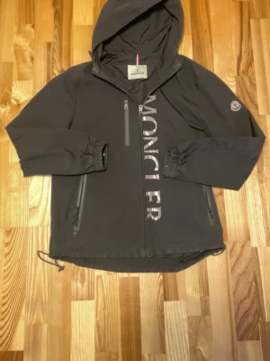 Svart vindjacka från Moncler - Snygg svart vindjacka från Moncler med huva och dragkedja framtill. Jackan har stora vita Moncler-bokstäver längs dragkedjan, två snedställda fickor med dragkedja och elastiska muddar vid ärmsluten. Materialet är tunt och vindtätt, perfekt för blåsiga dagar.