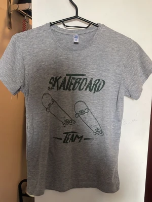 Grå t-shirt med skateboardtryck - Säljer en grå t-shirt från Fruit of the Loom med coolt skateboardmotiv och texten 'Skateboard Team' på framsidan. T-shirten har korta ärmar och är gjord i mjuk bomull, perfekt för dig som gillar skate och streetstyle.
