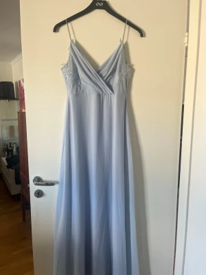 Ljusblå balklänning med spetsdetaljer - Elegant ljusblå långklänning med tunna axelband och omlott framtill. Klänningen har fina spetsdetaljer vid bysten och ett flytande, mjukt fall. Perfekt för speciella tillfällen där du vill känna dig extra fin.