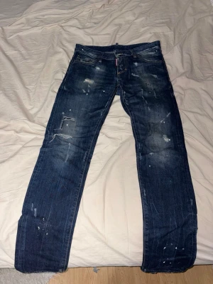 Dsquared2 jeans storlek 44 - Säljer ett par mörkblå jeans från Dsquared2 med coola slitningar, färgstänk och distressed detaljer. Klassisk femficksmodell med gul kontrastsöm och Dsquared2-logga på röd etikett vid gylfen. Perfekta för en trendig och avslappnad streetwear-look.