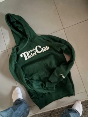 Grön hoodie St. Barth Padel Club - Mörkgrön hoodie från St. Barth med stor vit text 'Padel Club' på bröstet. Klassisk huva och magficka framtill. Tillverkad i mjukt material, perfekt för en avslappnad stil. Snygg och enkel design som sticker ut med trycket.