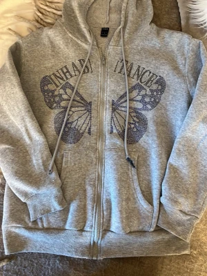 Grå hoodie med fjäril från SHEIN❣️ - Grå zip-hoodie från SHEIN med stor fjäril och texten 'INHABITANCE' i svart på bröstet. Hoodien har dragsko i huvan, fickor framtill och ribbade muddar. Tillverkad i mjukt sweatshirtmaterial, perfekt för en chill och avslappnad stil.