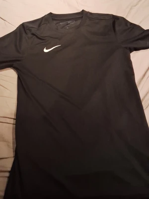 Svart Nike Dri-Fit träningströja - Säljer en svart träningströja från Nike med Dri-Fit-teknologi och slim fit-passform. Tröjan har rund halsringning och en vit Nike-logga på bröstet. Perfekt för träning och sportaktiviteter.