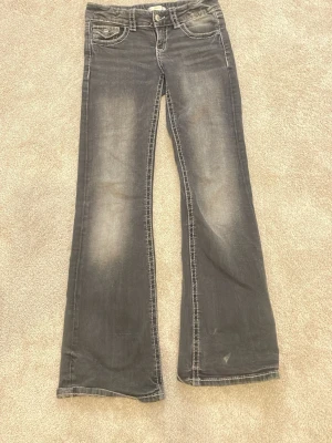 Svarta bootcut jeans med slitningar - Säljer ett par svarta bootcut jeans med snygga slitningar och kontrastsömmar. Jeansen har fem fickor, dekorativa knappar på bakfickorna och en klassisk låg midja. Perfekta för dig som gillar en avslappnad men trendig look. Storlek 152❤️❤️