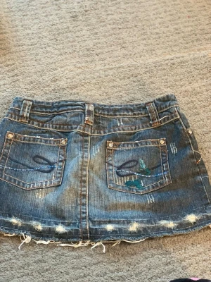 Blå jeanskjol med slitningar och broderi - Snygg blå jeanskjol i kort modell med slitna detaljer och rå kant nertill. Kjolen har två bakfickor med dekorativa broderier och små nitar. Perfekt för en avslappnad och trendig look.