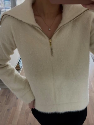 Krämvit stickad tröja med dragkedja - Mysig krämvit stickad tröja med halv dragkedja framtill och bred krage. Tröjan har lång ärm och är gjord i ett fluffigt, mjukt material som känns skönt mot huden. Perfekt för lager på lager och enkel att styla till olika outfits.