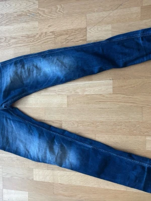 Blå slim fit jeans från Replay - Säljer ett par blå slim fit jeans från Replay med klassisk femficksdesign och snygga slitningar. Jeansen har en mörk tvätt och är tillverkade i bomull med lite stretch för extra komfort. Perfekta för en avslappnad och trendig look.