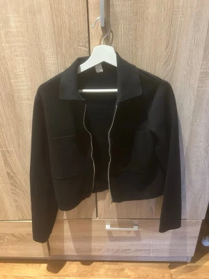 Svart zip cardigan med krage - Stilren svart cardiganjacka med dragkedja framtill och klassisk krage. Jackan har två stora fickor på framsidan och är tillverkad i ett mjukt stickat material. Perfekt för lager på lager och enkel att matcha med olika outfits.