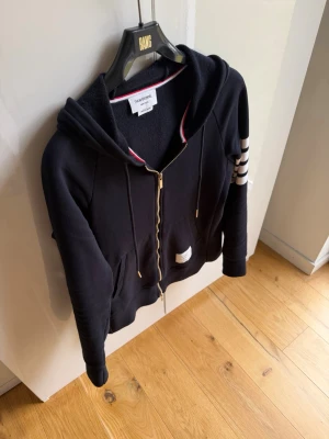 Marinblå hoodie från Thom Browne - Snygg svart hoodie från Thom Browne med dragkedja, huva och snörning. Klassiska vita ränder på ena ärmen och gulddetaljer på snörena. Röd, vit och blå kant på insidan av kragen ger en extra touch. Perfekt för en chill och stilren look. Påse samt tags medföljer! Endast tags avklippta. Mvh ItsForfits 🌟