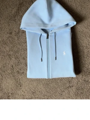 Ljusblå hoodie från Polo Ralph Lauren - Snygg ljusblå hoodie med dragkedja från Polo Ralph Lauren. Klassisk design med huva, dragsnören och fickor framtill. Ikoniska vita loggan broderad på bröstet. Perfekt för en chill och stilren look.