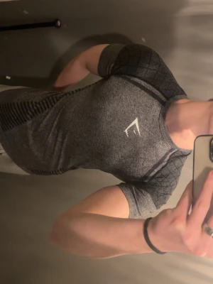 OG Gymshark - Säljer en tight grå träningströja från Gymshark med svarta detaljer och korta ärmar. Tröjan har ett sportigt mönster längs sidorna och axlarna samt Gymsharks logga på bröstet. Perfekt för gymmet eller löpning.