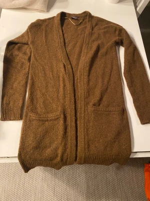 Brun lång kofta från ICHI - Brun lång kofta från ICHI med öppen front och två fickor framtill. Koftan är stickad i ett mjukt material och har ribbade muddar vid ärmslut och nederkant. Använd fåtal gånger.