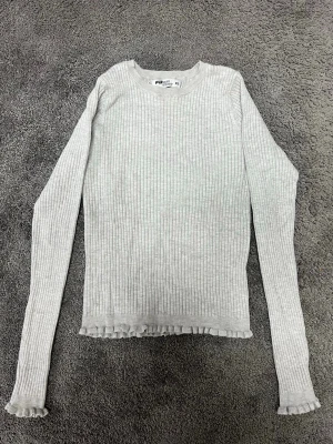 Ljusgrå ribbad långärmad topp - Superfin ljusgrå ribbad topp med långa ärmar och volangkant nertill och vid ärmsluten. Perfekt att styla med jeans eller kjol för en chill look.