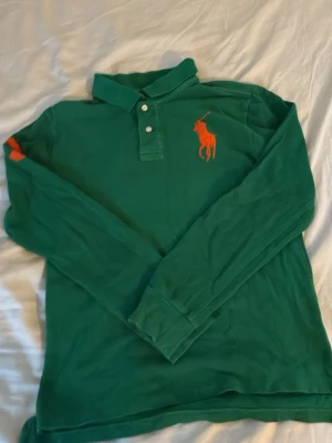 Grön långärmad skjorta från Polo Ralph Lauren - Säljer en grön långärmad skjorta från Polo Ralph Lauren med orange broderad logga på bröstet och en stor orange bokstav på ärmen. Klassisk krage och knappar framtill. Perfekt för dig som gillar sportig och stilren look.