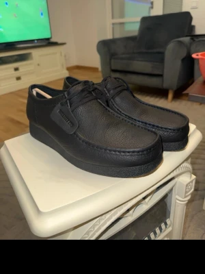 Svarta Wallabee Evo loafers - Säljer ett par svarta Wallabee Evo loafers från Clarks. Aldrig använda. Skorna är tillverkade i slätt, strukturerat läder och har klassisk mockasinkant samt snörning framtill. Tidlös design med robust sula och diskret tag på sidan.