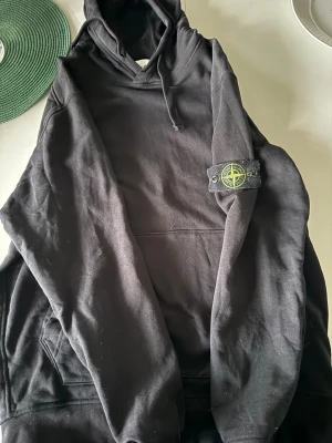 Svart hoodie från Stone Island - Säljer en svart hoodie från Stone Island med klassisk logotyp-patch på ärmen. Hoodien har huva med dragsko och en stor magficka framtill. Tillverkad i mjukt bomullsmaterial som är skönt att bära. Perfekt för dig som gillar streetwear och vill ha en stilren look.