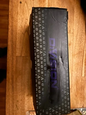 50 sport cylle  - Säljer en svart kartong från Division med tydlig logga i lila och vitt. Kartongen har ett grafiskt mönster med texten 'DIVISION' tryckt runt om. Perfekt för förvaring eller som presentlåda. Materialet är kartong och designen är modern och stilren.