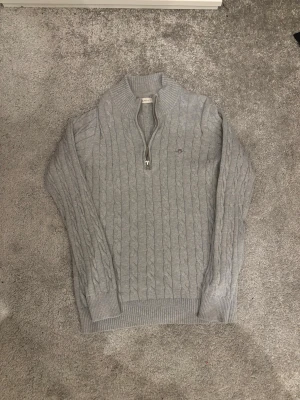 Grå kabelstickad tröja från GANT - Snygg grå kabelstickad tröja från GANT med halv dragkedja och hög krage. Tröjan har ribbade muddar och ett litet broderat GANT-logo på bröstet. Perfekt för lager-på-lager och passar till det mesta.