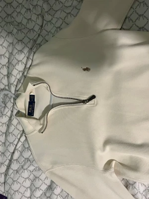 Vit halvzip tröja från Polo Ralph Lauren - Snygg vit tröja med halvzip och klassisk Polo Ralph Lauren-logga broderad på bröstet. Tröjan har hög krage och är tillverkad i ett mjukt material som känns skönt mot huden. Perfekt för att ge din outfit en clean och stilren look.
