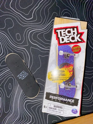 Tech Deck fingerboard Santa Cruz - Fingerboard från Tech Deck Performance Series med Santa Cruz-design. Brädan har blått mönster och Santa Cruz-logga i rött och gult med eldflamma. Kommer med vita hjul och silverfärgade trucks. Perfekt för tricks och samling.