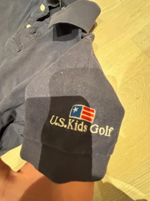 Blå pikétröja från U.S. Kids Golf - Snygg blå pikétröja från U.S. Kids Golf med broderad logga och amerikansk flagga på ärmen. Tillverkad i 100% bomull och har klassisk krage samt knappar framtill. Perfekt för en sportig och avslappnad stil.