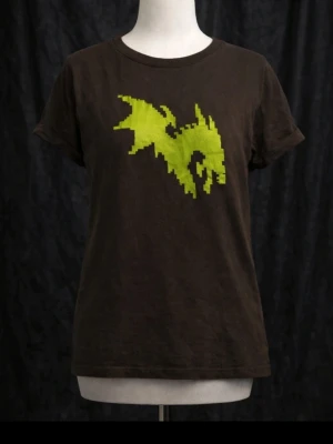 Grön T‑shirt med lime färgadpixel‑drake – retro & nördig - En mörkgrön T‑shirt med ett starkt grönt pixelmotiv av en drakliknande varelse mitt på bröstet från När on. Designen har en tydlig retro‑gaming‑känsla med blockiga former och fantasy‑touch  perfekt för dig som gillar pixelart, spelestetik eller bara vill ha ett statement‑plagg som sticker ut. Mjuk och bekväm passform som funkar både till vardags och event.