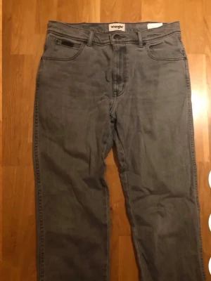 Grå jeans från Wrangler - Säljer ett par klassiska grå jeans från Wrangler med rak passform och fem fickor. Jeansen har bälteshällor, knappgylf och Wrangler-logga på bakfickan. Tillverkade i slitstark denim som passar perfekt till en avslappnad stil.