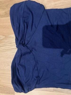 Mörkblå ribbad bandeau topp - Snygg mörkblå bandeau topp med ribbad struktur och twistad detalj framtill. Toppen är axelbandslös och har en croppad passform som passar perfekt till höga jeans eller kjol. Enkel att styla för en trendig look.