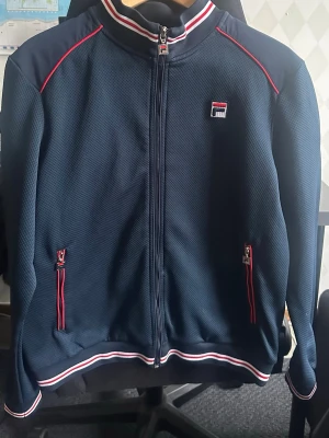 Fila Kofta  - Snygg marinblå ziptröja från Fila med rödvita detaljer på krage, muddar och dragkedjor. Tröjan har två fickor med dragkedja och ett Fila-märke på bröstet. Sport material, skön passform, gott skick. STORLEK L men passar M också 