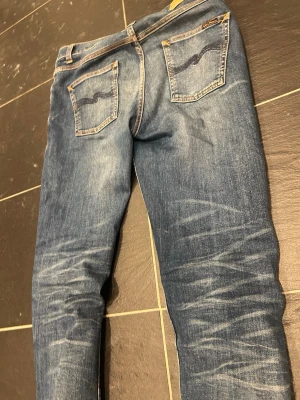 Blå fit jeans från Nudie Jeans - Snygga blå slim fit jeans från Nudie Jeans med klassisk femficksdesign och tydliga slitningar framtill och på baksidan. Jeansen har kontrastsömmar och Nudie-logga på bakfickorna. Tillverkade i denim med stretch för extra komfort. Storlek 31L32, modell: lean dean. Sjuk wash 