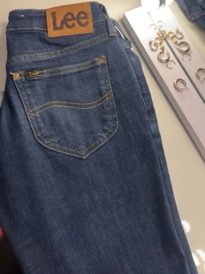 Blå jeans från Lee med klassisk passform - Säljer ett par blå bootcut jeans från Lee med klassisk femficksdesign och snygga kontrastsömmar. Jeansen har en normal midja och raka ben, tillverkade i slitstarkt denim-material. Perfekta för dig som gillar tidlös stil och vill ha ett par jeans som funkar till allt. Använda runt 4-5 gånger. Köpta för 1099kr. Strl 24 längd 31