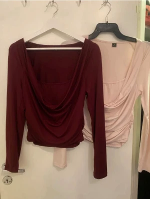 Vinröd draperad topp med lång ärm - Trendig vinröd topp med draperad urringning och långa ärmar. Modellen är figurnära och croppad, vilket ger en snygg siluett. Tillverkad i mjukt stretchigt material som känns bekvämt mot huden. Perfekt för dig som vill ha en stilren men ändå unik look.