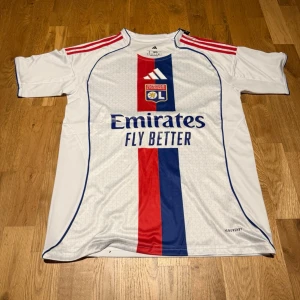 Lyon home kit 25/26 X endrick 9 - Snygg vit Olympique Lyonnais matchtröja från Adidas med röda och blå detaljer, klubbmärke på bröstet och sponsortryck 'Emirates Fly Better'. Röd och blå rand framtill, namn och nummer 'Endrick 9' på ryggen. Perfekt för fotbollsfans!