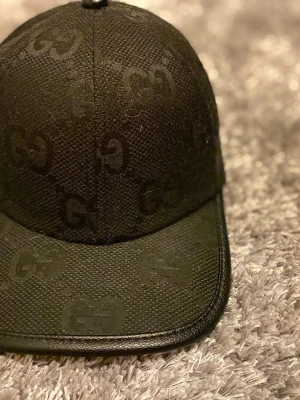 Svart keps från Gucci med GG-mönster - Snygg svart keps från Gucci med det klassiska GG-monogrammet över hela kepsen. Tillverkad i ett tåligt canvasmaterial med böjd skärm och justerbar baksida. Perfekt accessoar för en trendig och exklusiv look. Skickar samma dag eller dagen efter, är villig att sänka priset vid snabb o smidig affär mvh☺️