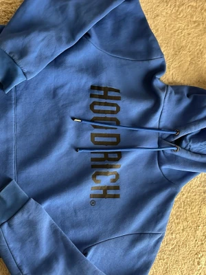 Blå HOODRICH hoodie med tryck - Snygg blå hoodie från HOODRICH med svart tryck på bröstet. Klassisk modell med huva och dragsnören, ribbade muddar vid ärmar och midja. Tillverkad i mjukt bomullsmaterial som känns skönt mot huden. Perfekt för en avslappnad streetwear-look. Slitade snören, använd.