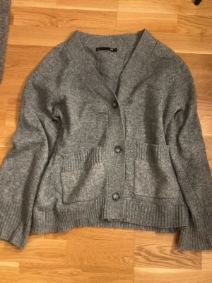 Grå kofta med fickor och knappar - Mysig grå kofta med v-ringning och tre knappar framtill. Två stora ribbstickade fickor och breda muddar vid ärmslut och nederkant. Perfekt för lager på lager och enkel att matcha med olika outfits.