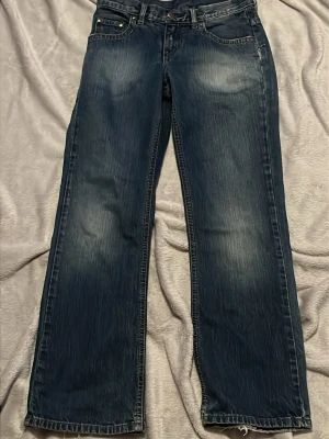 Mörkblå bootcut/straight jeans från dgate - Snygga mörkblå bootcut jeans från dgate med klassisk femficksdesign och slitningar framtill och baktill. Jeansen har normal midja och raka ben med lätt utsvängd passform. Tillverkade i denim med tydliga kontrastsömmar och läderpatch bak i midjan.