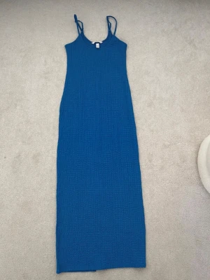 Blå långklänning med tunna band - En enkel och stilren blå långklänning med tunna axelband och rak passform. Klänningen har en lätt strukturerad yta och är perfekt för varma dagar. Materialet känns mjukt och luftigt, vilket gör den skön att bära.